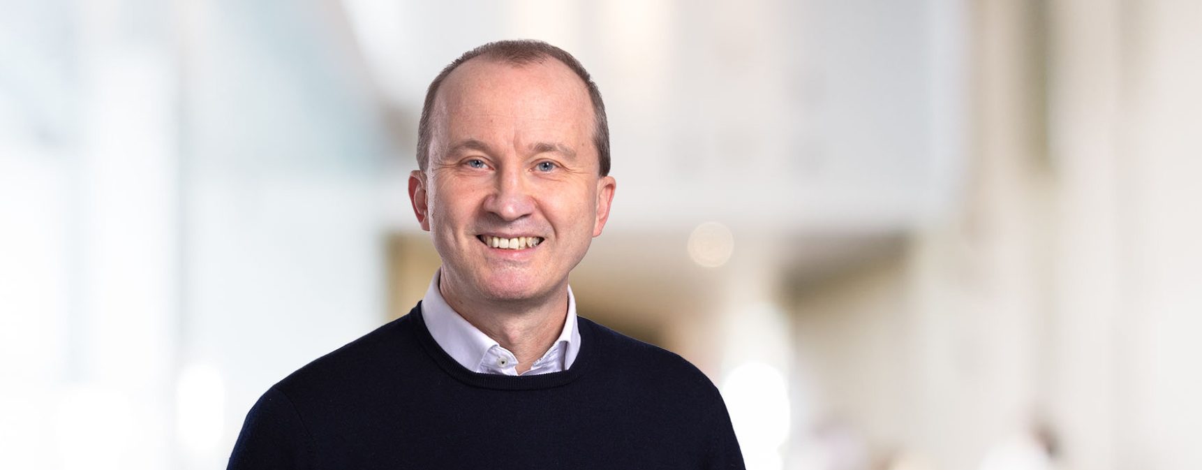 Hartmut Reiners in dunklem Pullover lächelt vor einem hellen, neutralen Hintergrund.